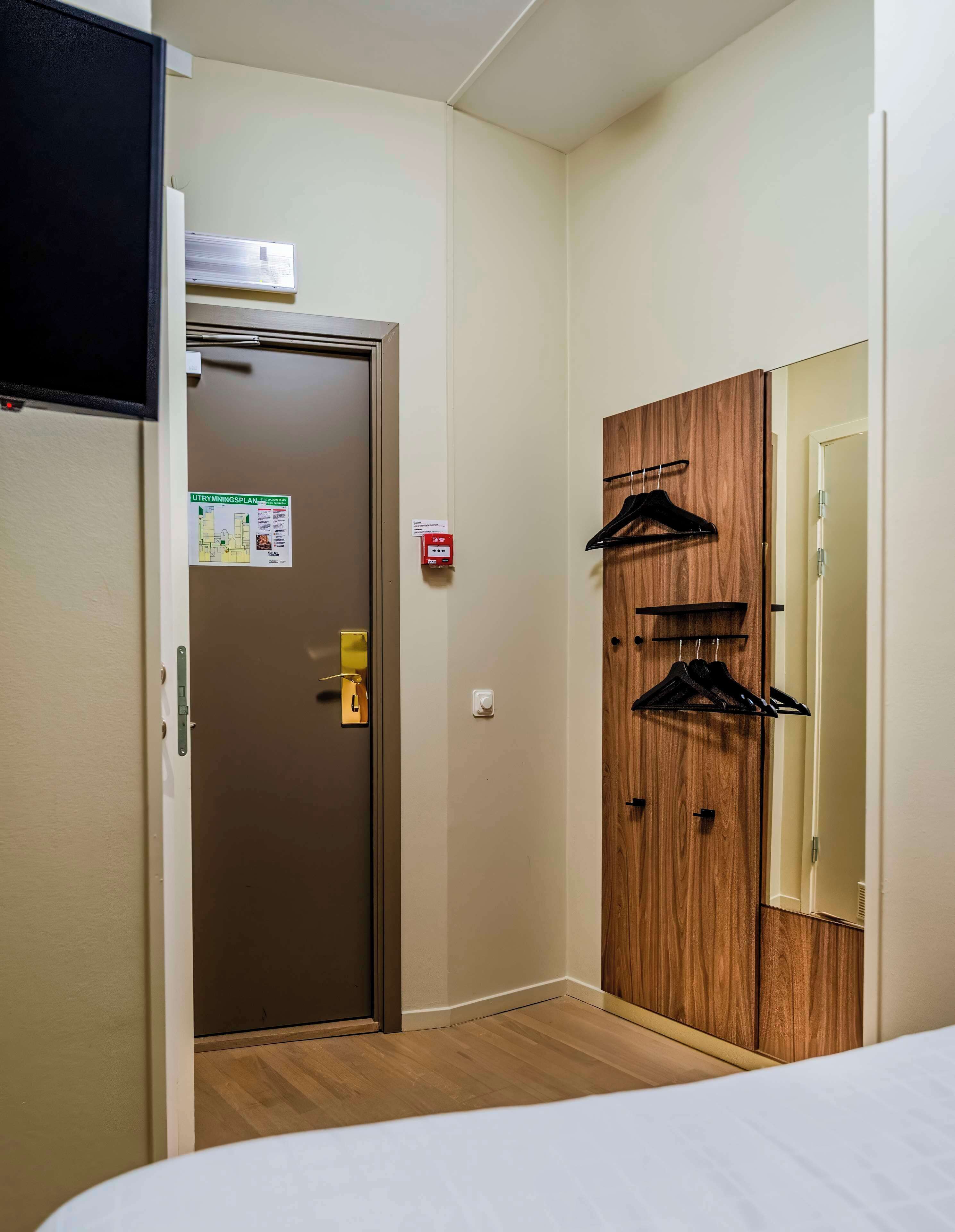 Ein Hotelzimmer mit Bett, einem Holzschrank zur Aufbewahrung von Kleidung, einer Tür mit Rauchmelder und einer Wand-Klimaanlage.