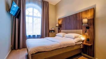 A neatly made bed with a headboard, a flat-screen TV mounted on the wall, and a window with curtains.