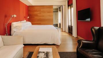 Une chambre d’hôtel avec un lit, un canapé, un fauteuil en cuir, un téléviseur à écran plat et un plancher en bois.
