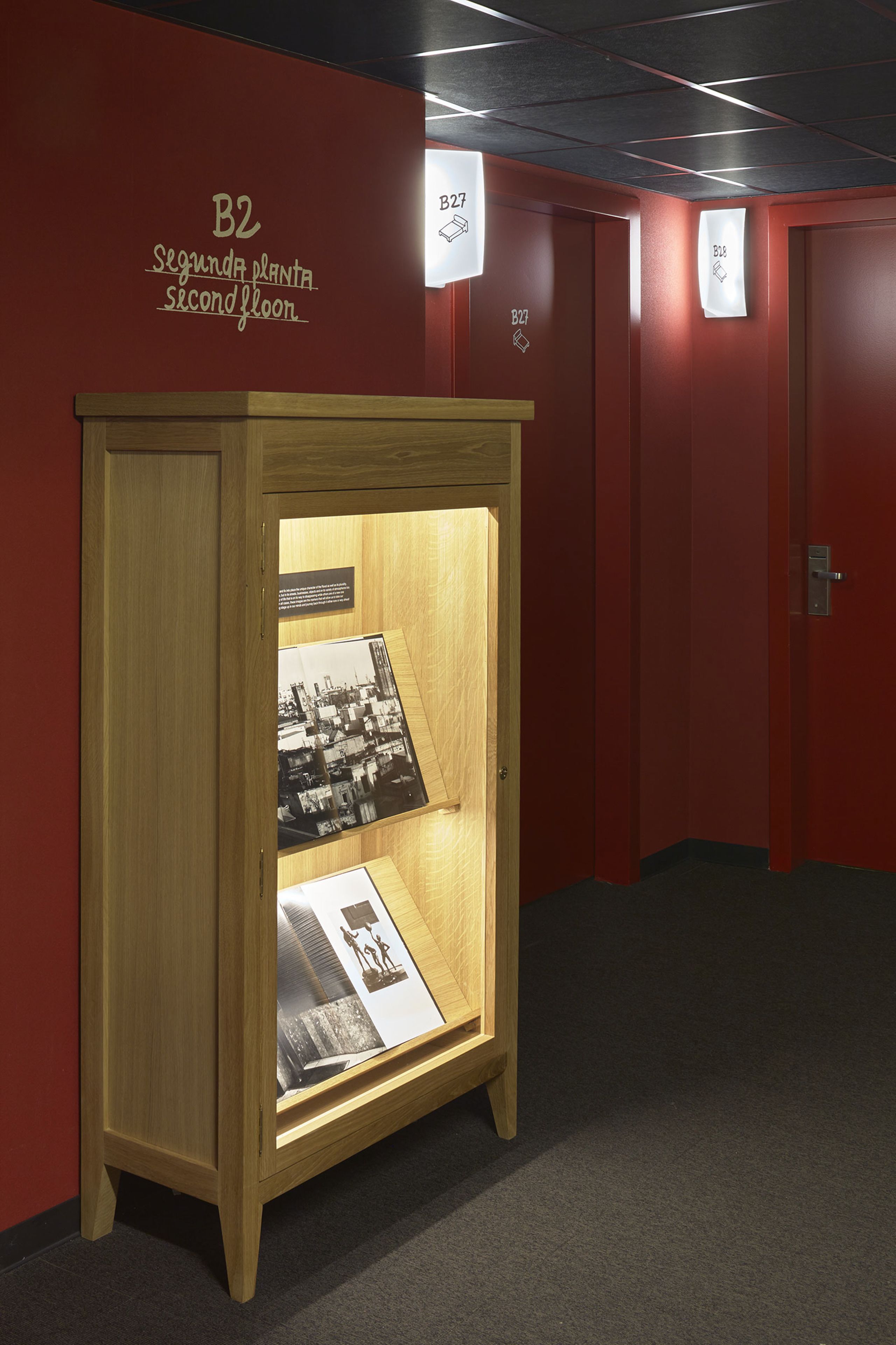 Un mueble de madera expositor con paneles informativos en un pasillo con paredes y puertas rojas, identificadas con B2 y B27.