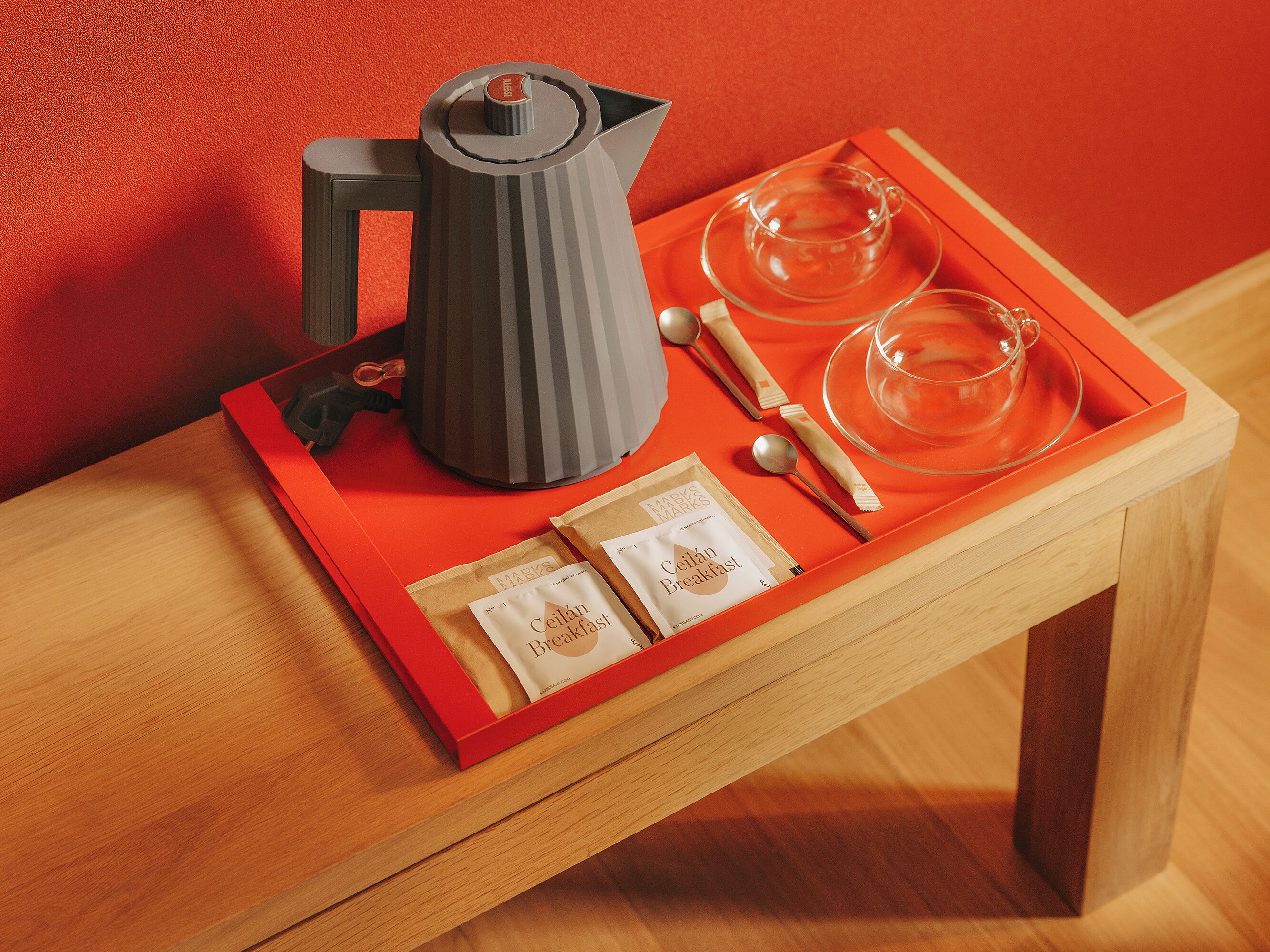 A tray with a coffee maker, teacups, and breakfast items on a wooden table against a red wall.