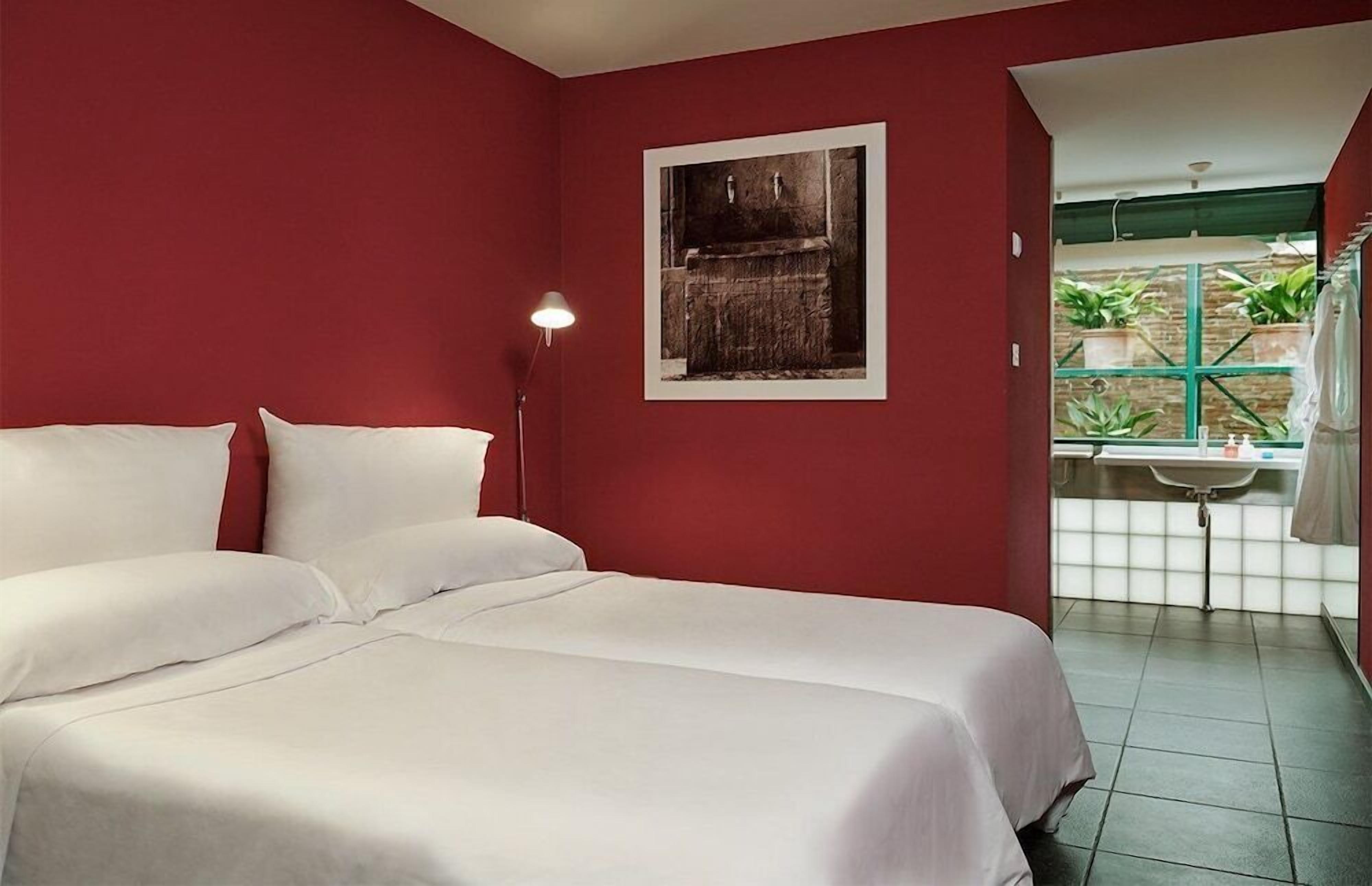 Una camera d'albergo con un letto, un comodino, un quadro appeso alla parete e una finestra con piante all'esterno.