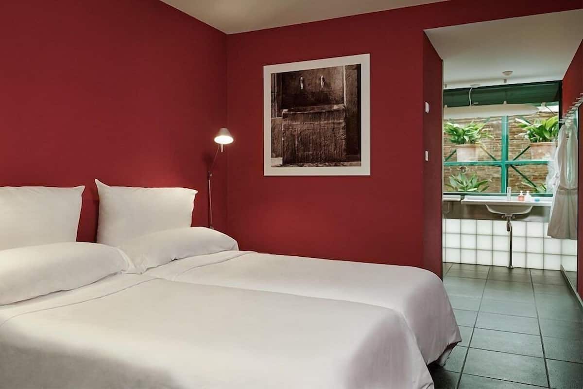 Ein Hotelzimmer mit einem Bett, einem Nachttisch, einem gerahmten Bild an der Wand und einem Fenster mit Pflanzen davor.