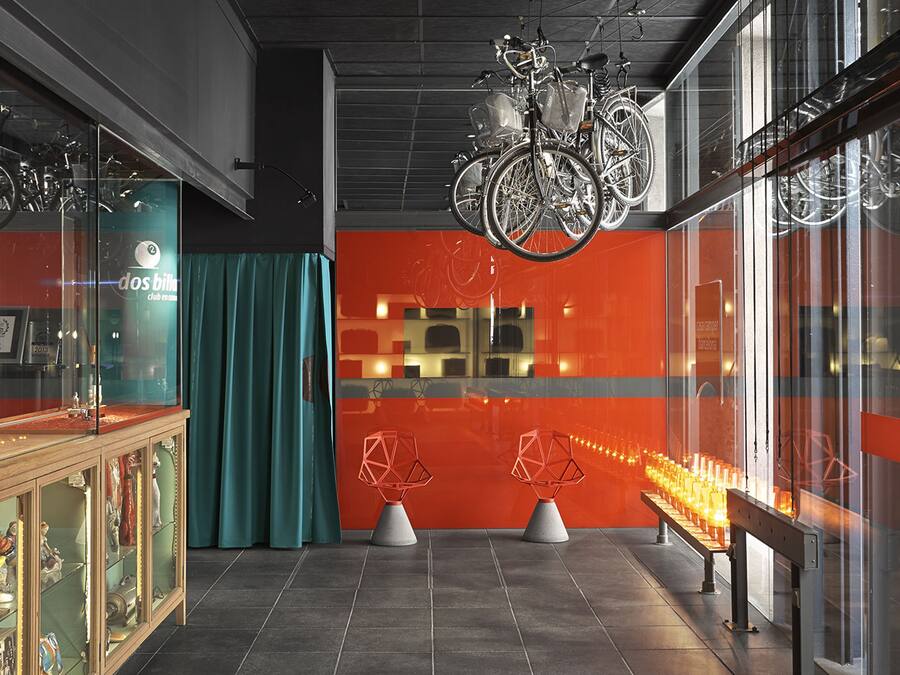 Un interior moderno con bicicletas colgando del techo, una vitrina de cristal con varios objetos y una zona de estar con un banco y dos sillas de diseño geométrico.