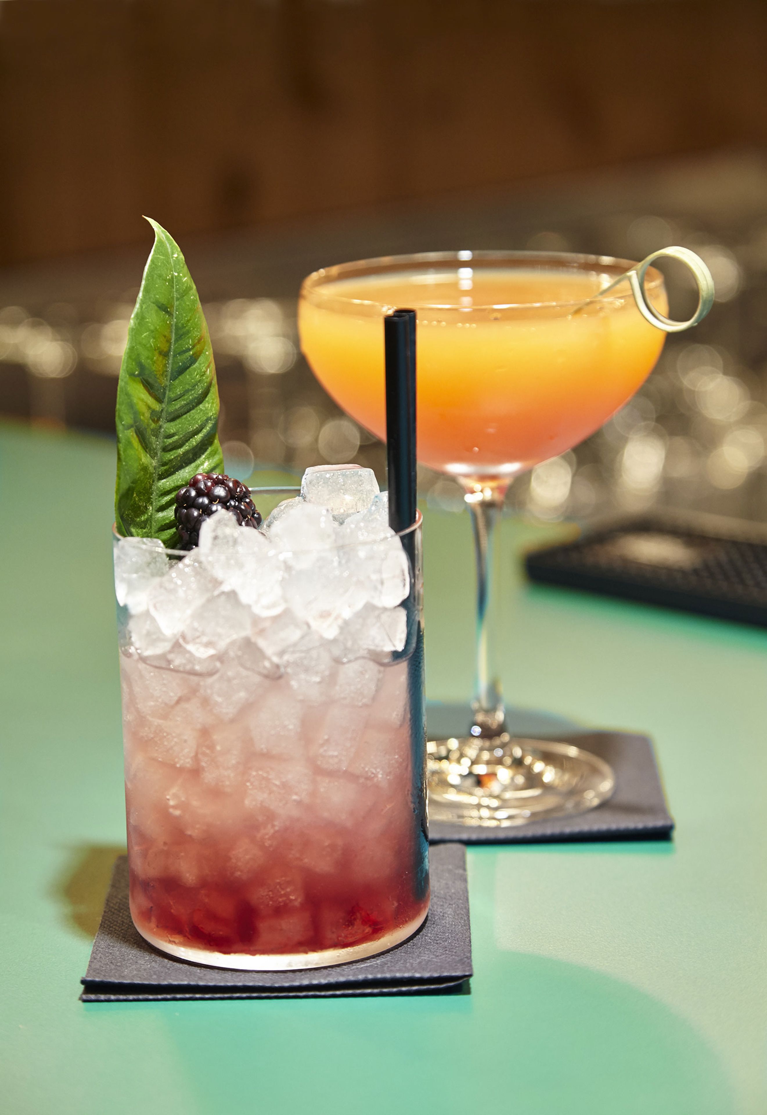 Two drinks on a table, one with ice and a blackberry, and the other with a straw.