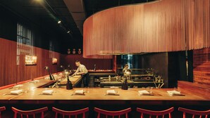 Un'area bar con un lungo bancone, sedie rosse e un barista che prepara cocktail.