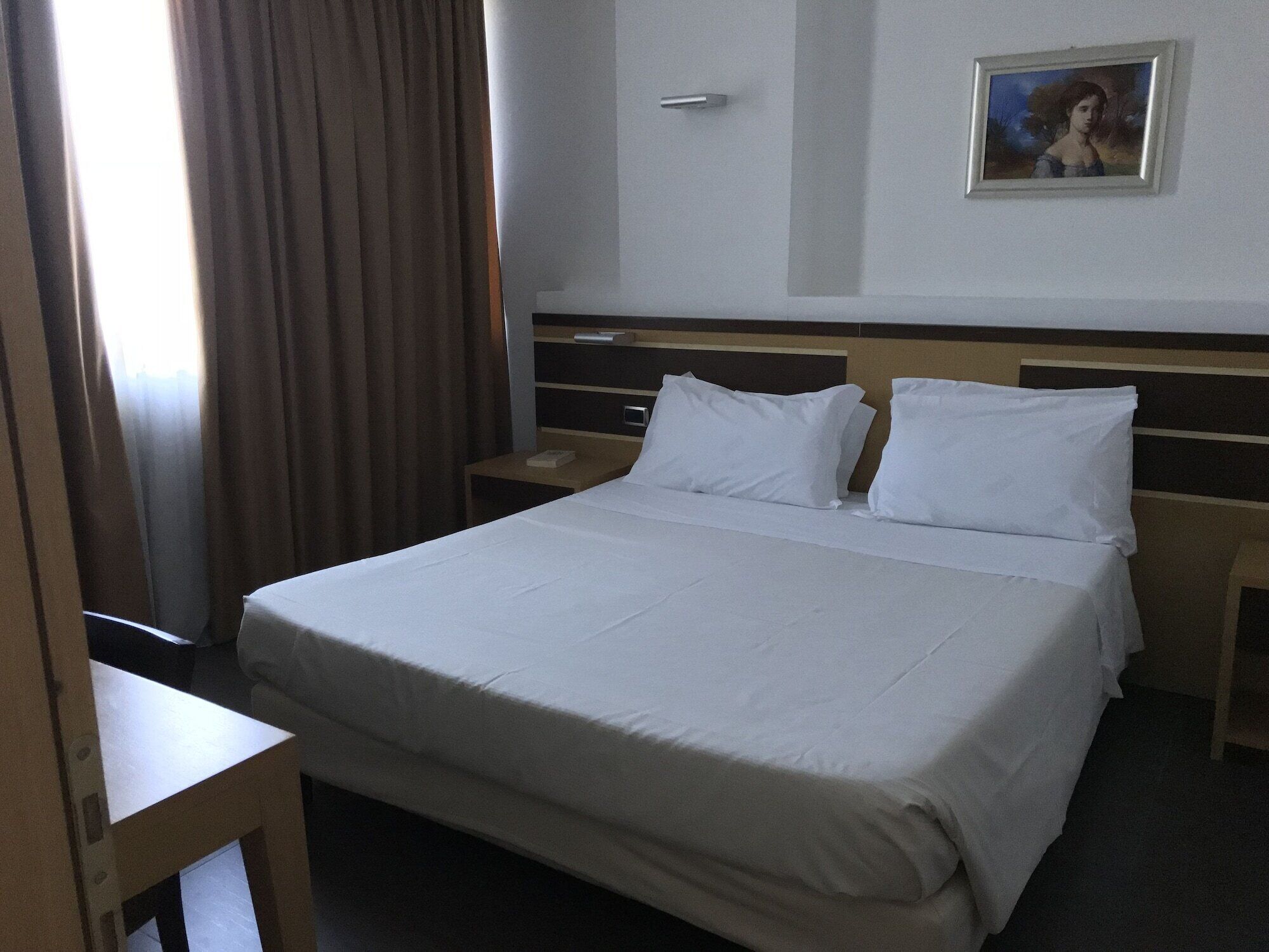 A hotel room with a large bed, two pillows, a nightstand, a bedside table, a wall-mounted painting, and a window with curtains.