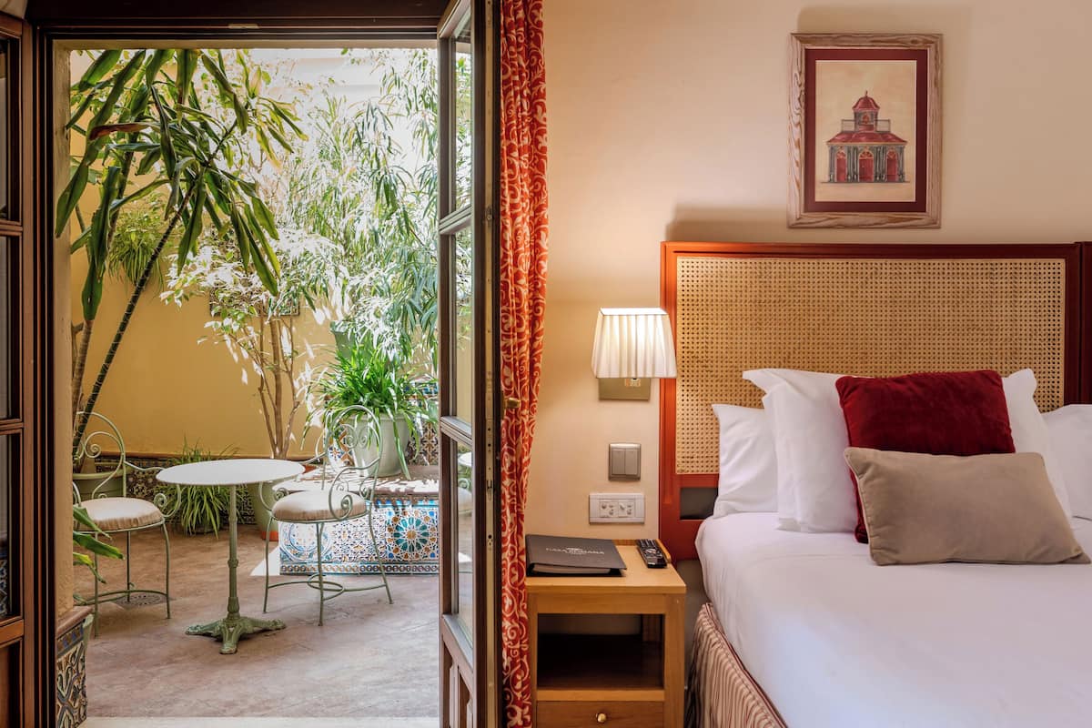 Een hotelkamer met een bed, nachtkastje, lamp en uitzicht op een terras met planten en een tafel.