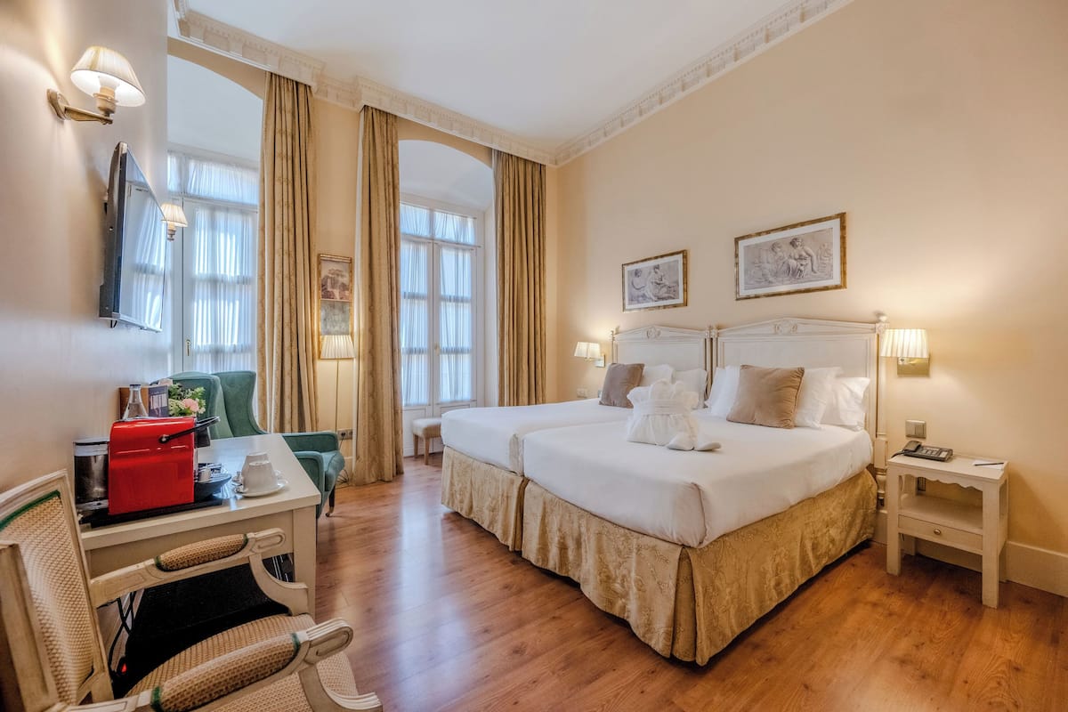 Een hotelkamer met een tweepersoonsbed, een bureau met een rood apparaat, een stoel, een nachtkastje en een raam met gordijnen.