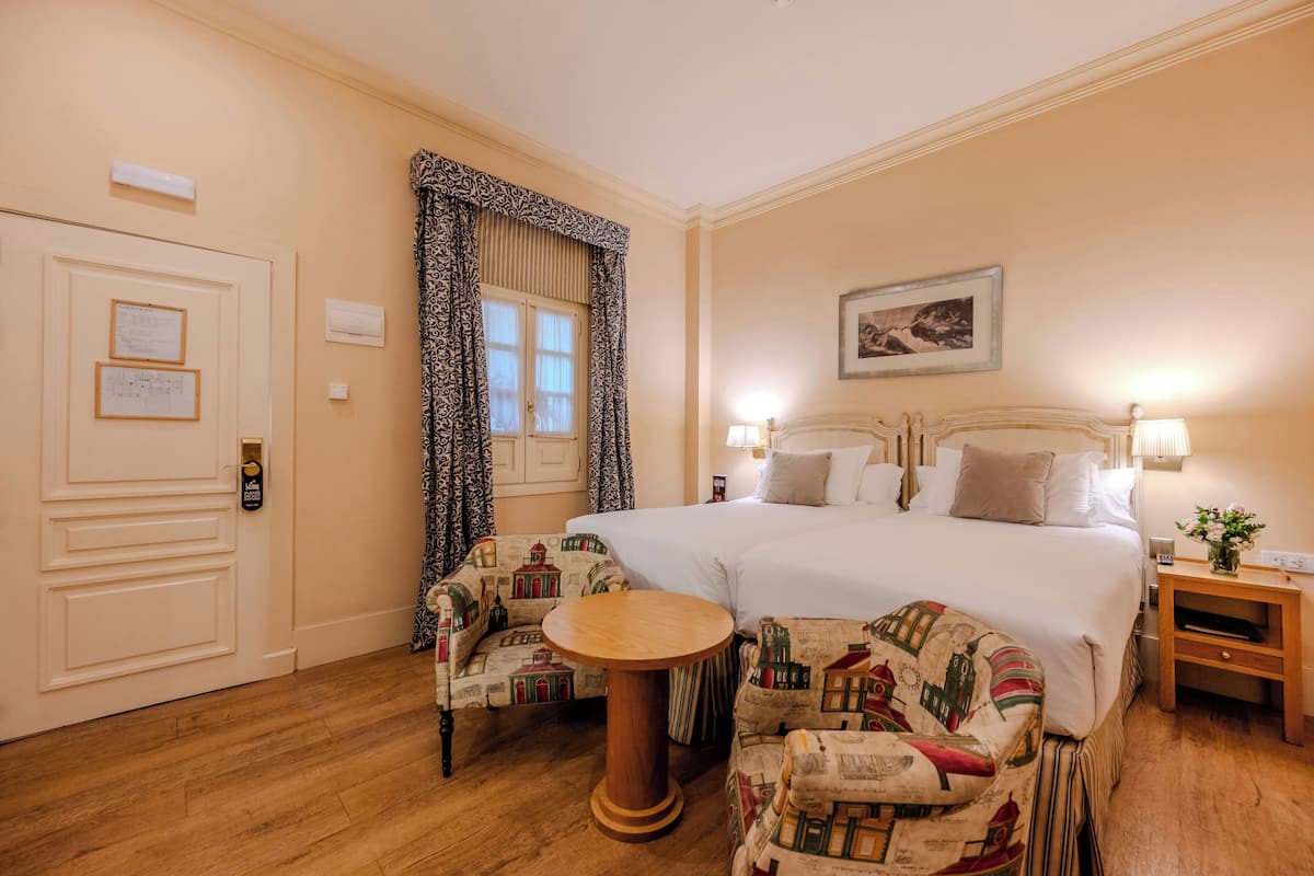 Hotelkamer met een groot bed, twee bedlampen, een klein rond tafeltje, gestoffeerde fauteuils, een houten vloer, een deur met slot en een ingelijst kunstwerk aan de muur.