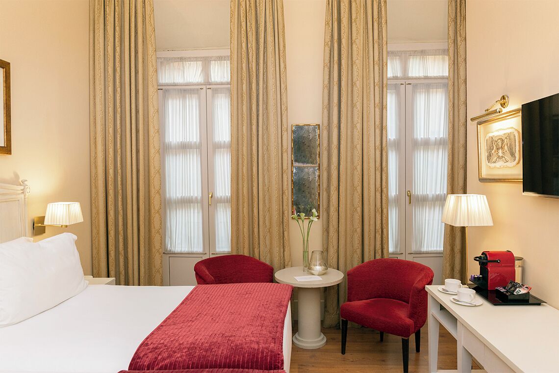 Een hotelkamer met een bed, twee rode stoelen, een kleine tafel met een vaas, een koffieapparaat en een televisie aan de muur.