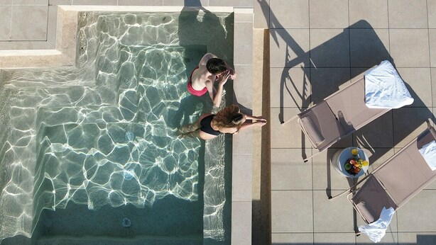 Une vue aérienne d’une piscine à l’eau claire, entourée d’un sol carrelé et agrémentée d’une table sur laquelle se trouvent une boisson et des fruits.