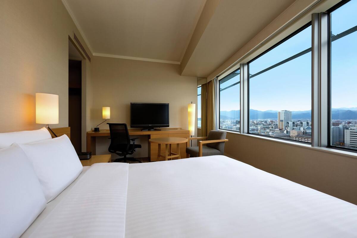 Une chambre d’hôtel avec un grand lit, un bureau avec une télévision, une chaise, une petite table et une vue sur le paysage urbain grâce à de grandes fenêtres.