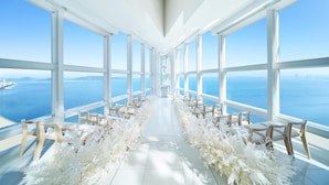 A high-ceilinged room with large windows, a white floor, and a central aisle with chairs.