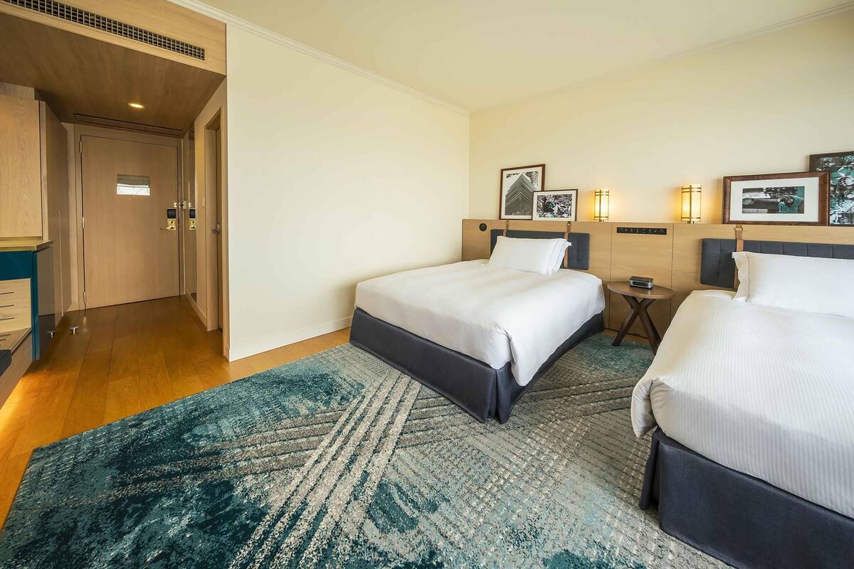 Une chambre d’hôtel avec deux lits, une porte en bois, une petite table de chevet et des lampes fixées au mur.
