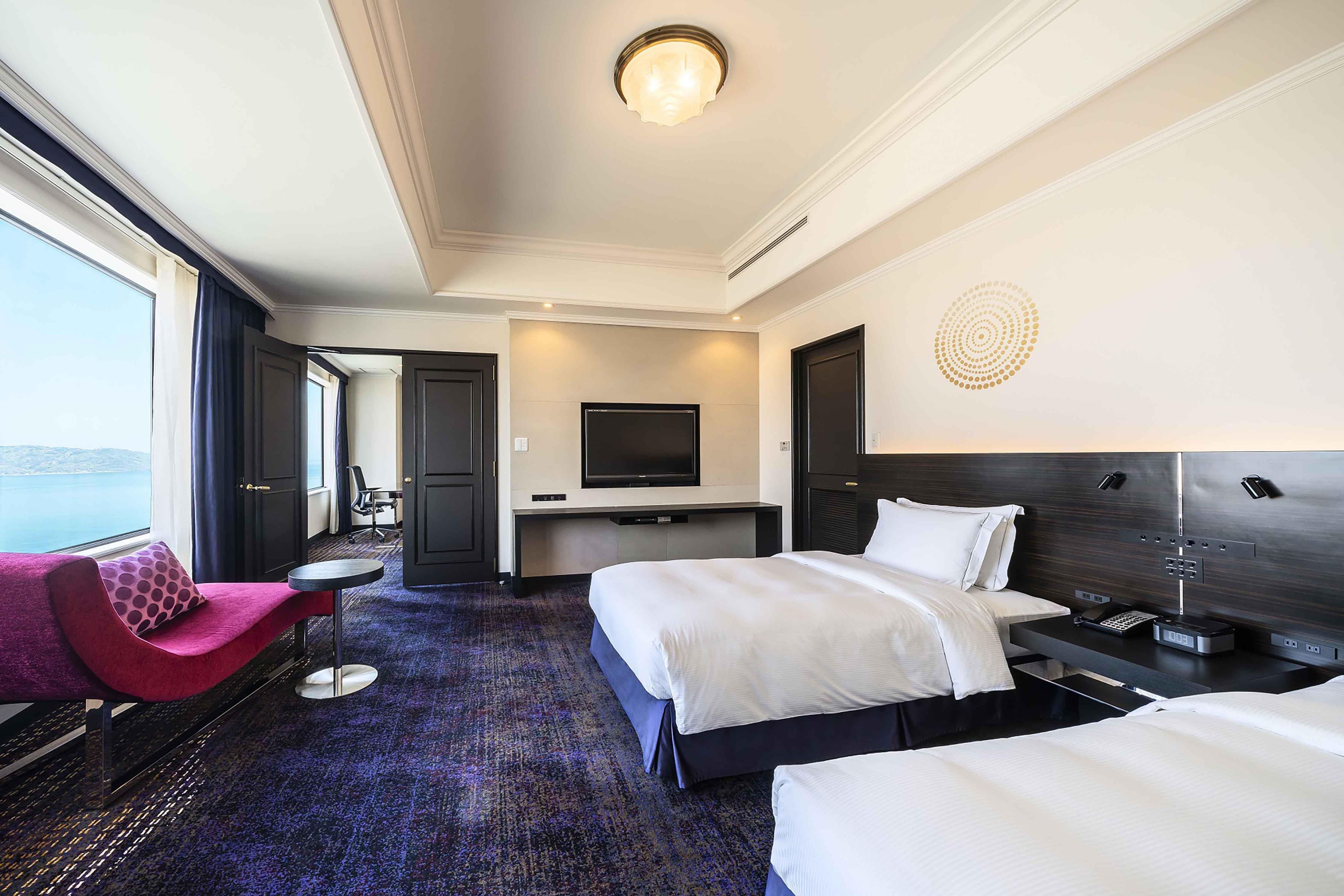 Une chambre d’hôtel avec un grand lit, un lit plus petit, un bureau avec un téléviseur à écran plat, une chaise et une vue sur la mer par une grande fenêtre.