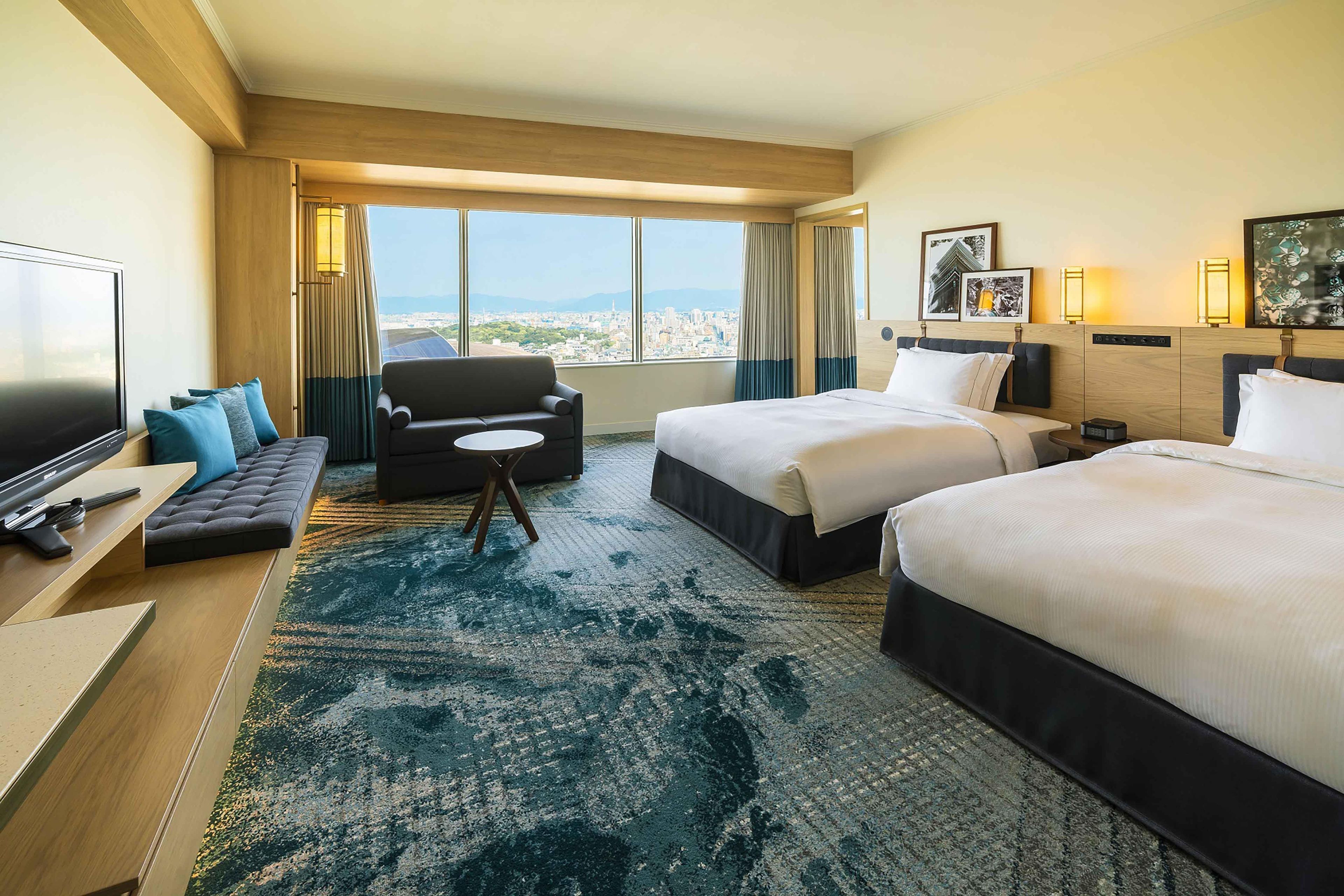 A hotel room with a large bed, a smaller bed, a sofa, a TV, and a view of a cityscape.