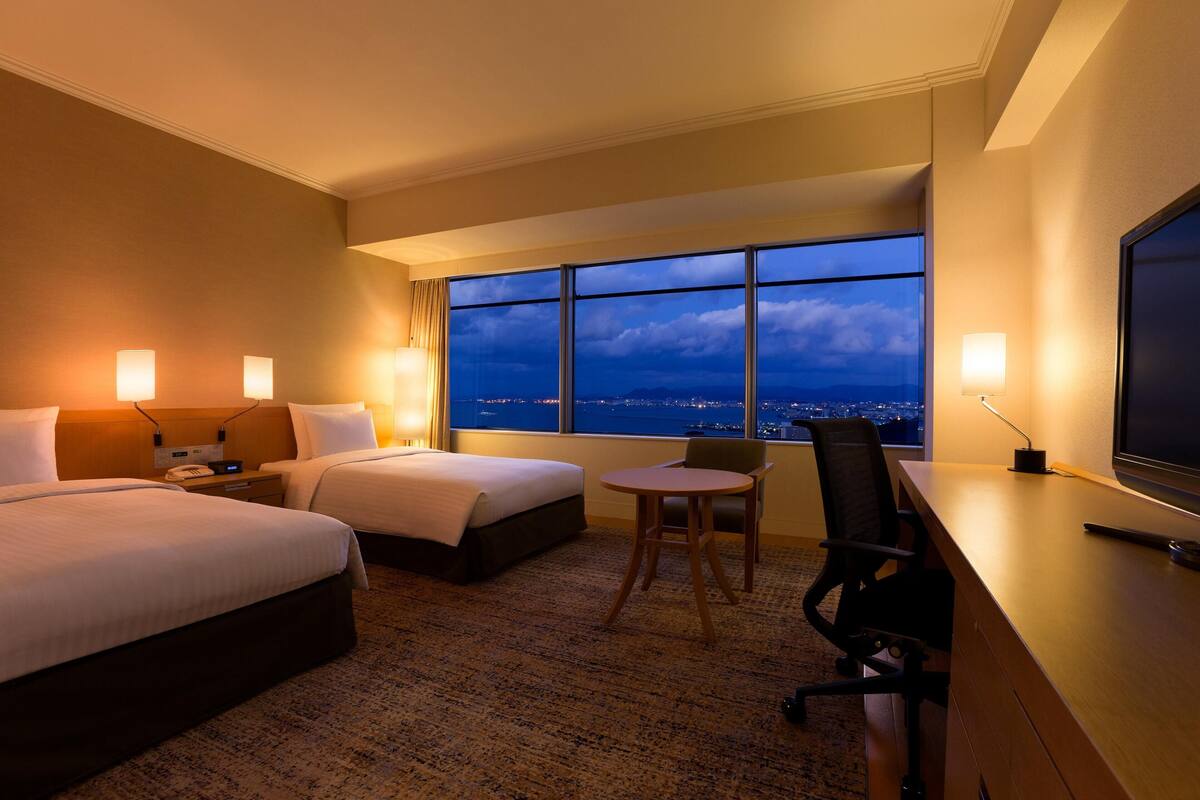 Une chambre d’hôtel avec deux lits, un bureau, une chaise, une télévision et une grande fenêtre donnant sur la ville.