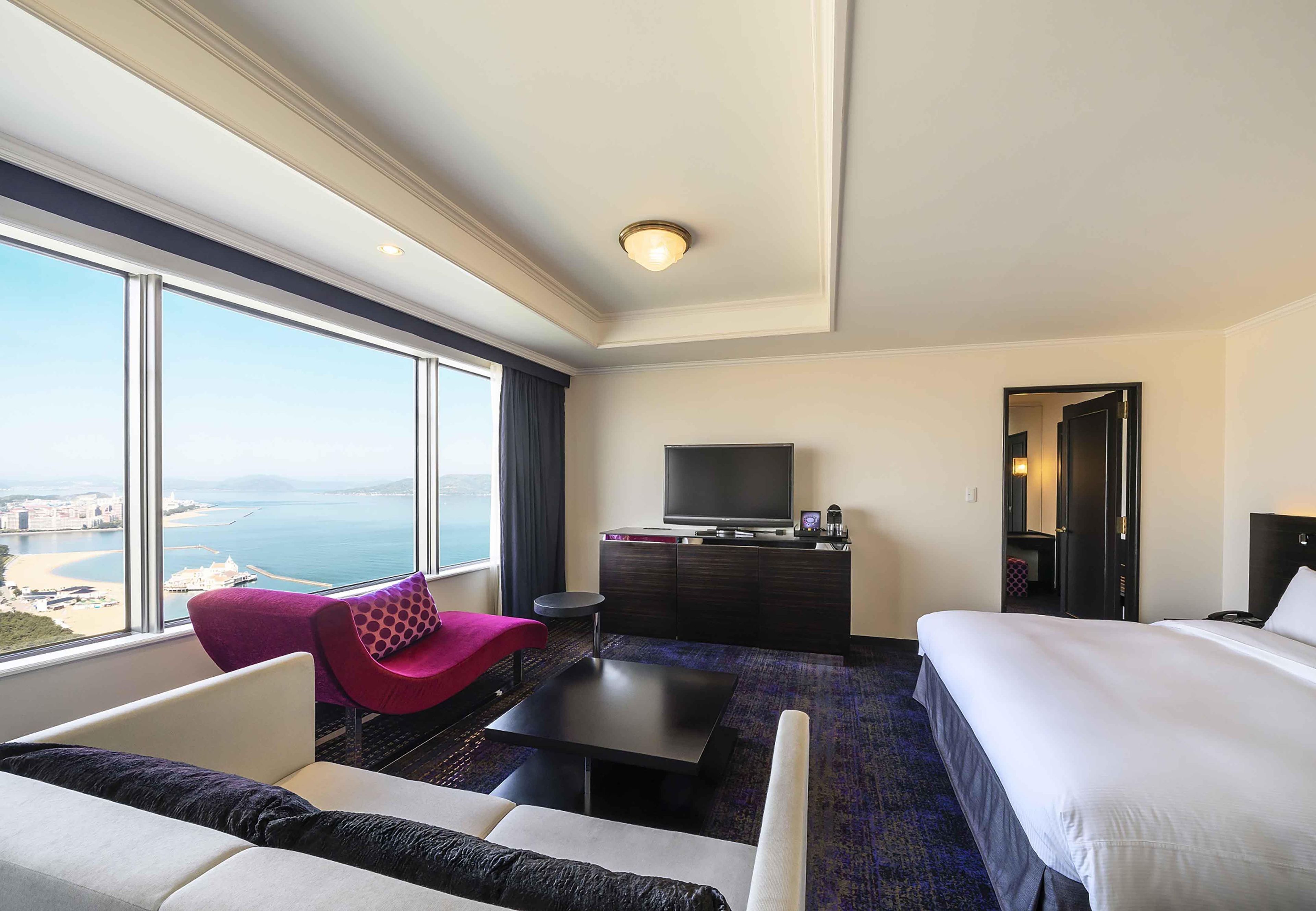 A hotel room with a large window, a bed, a sofa, a TV, and a small table.