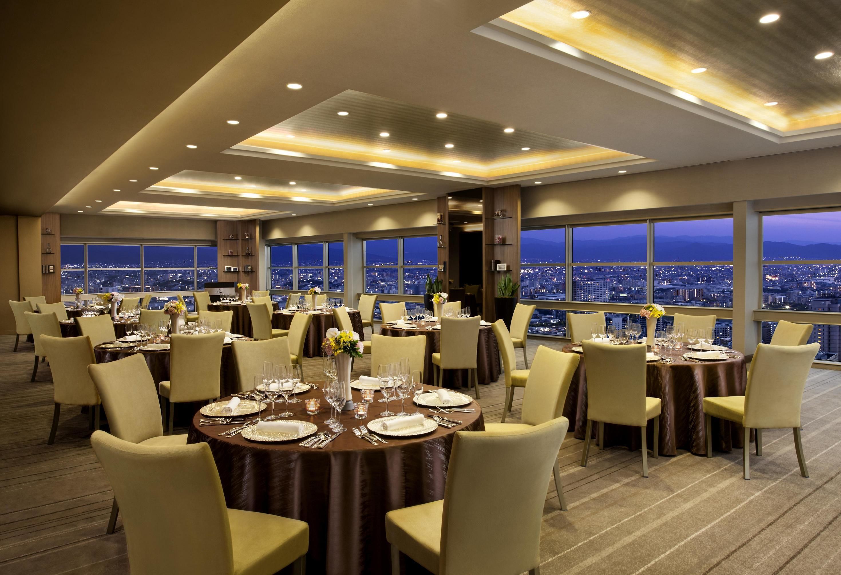 Un restaurant situé dans un hôtel de grande hauteur, avec de nombreuses tables rondes dressées pour le dîner, de grandes fenêtres offrant une vue sur la ville et un design moderne au niveau du plafond.