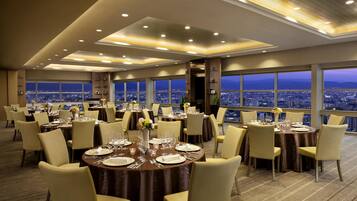A high-rise hotel dining area with multiple round tables set for dining, large windows offering city views, and a modern ceiling design.
