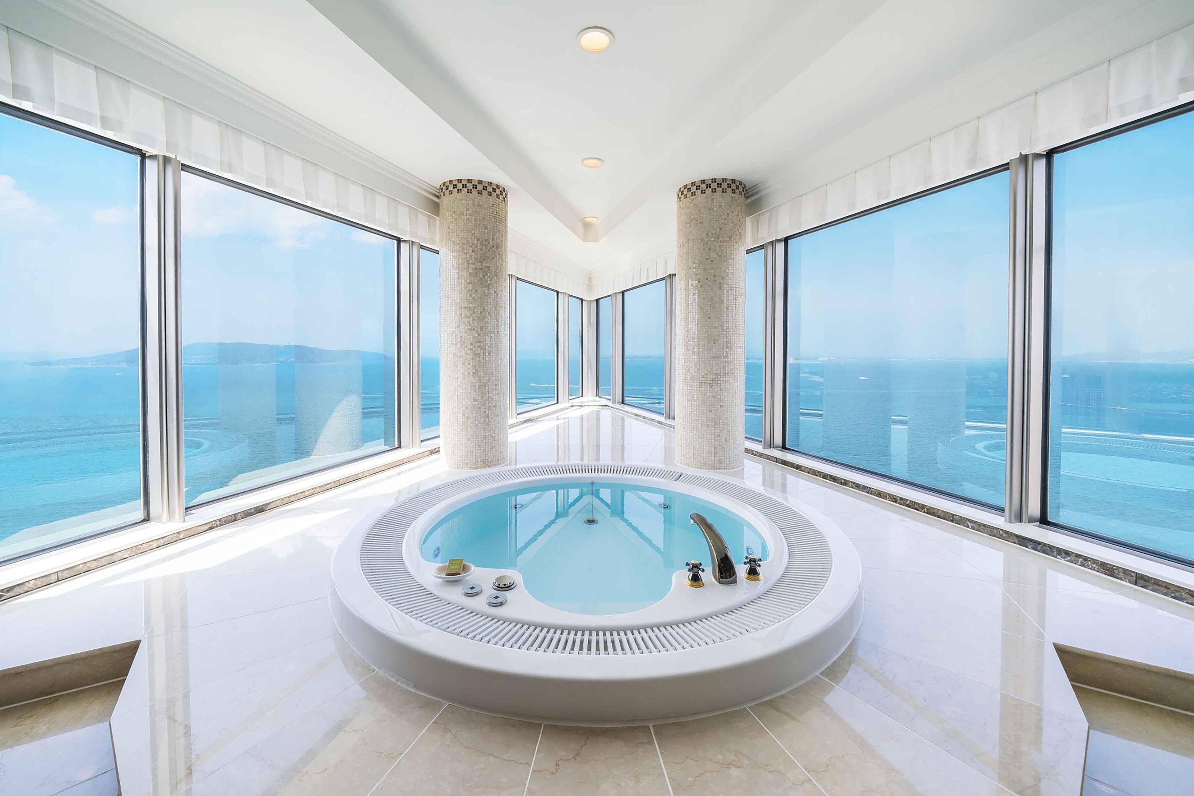 A modern indoor swimming pool with a panoramic ocean view, surrounded by large windows and minimalist columns.