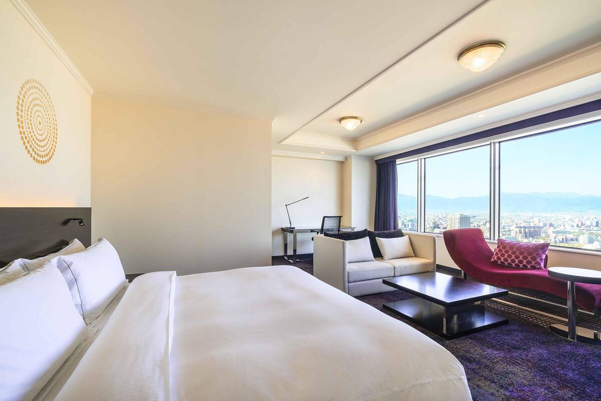 Une chambre d’hôtel avec un grand lit, un bureau, une chaise rouge et une vue sur la ville.