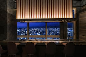 A bar with a city view at night, featuring a modern design with a large window and a slatted ceiling.