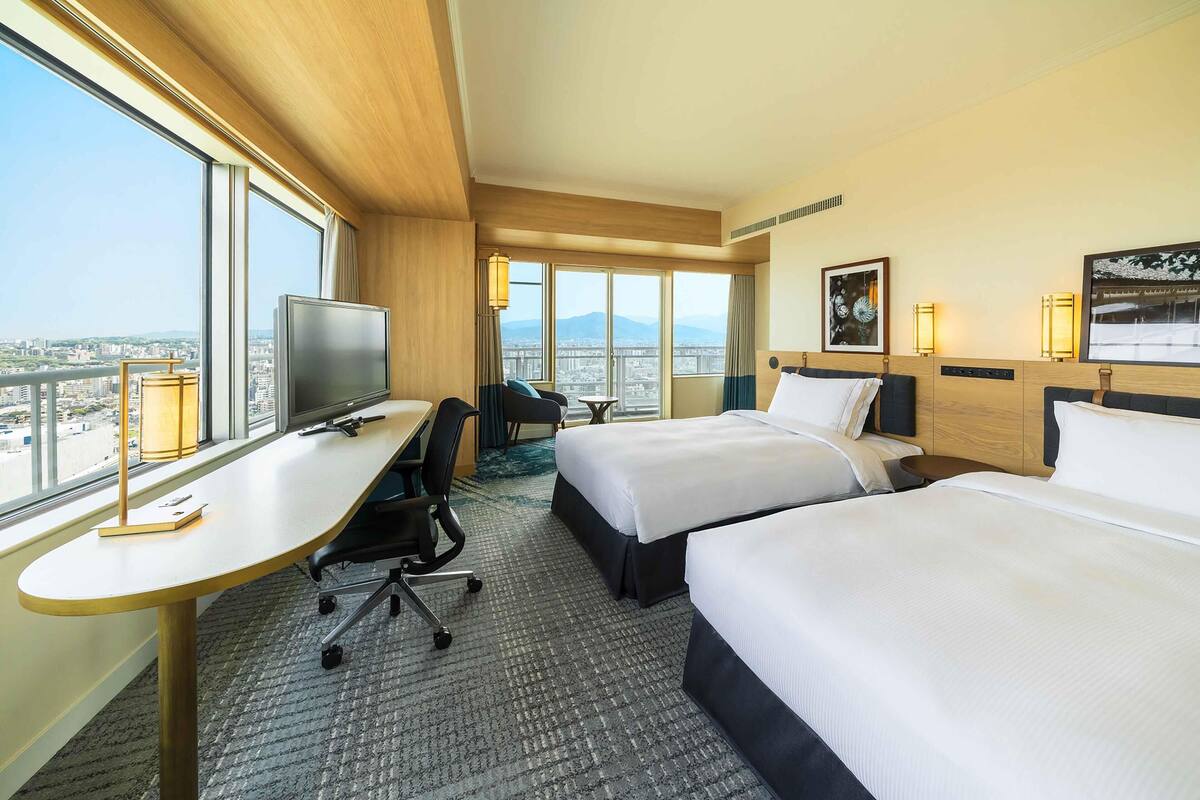 Une chambre d’hôtel avec deux lits, un bureau, une chaise, une télévision et de grandes fenêtres donnant sur la ville.