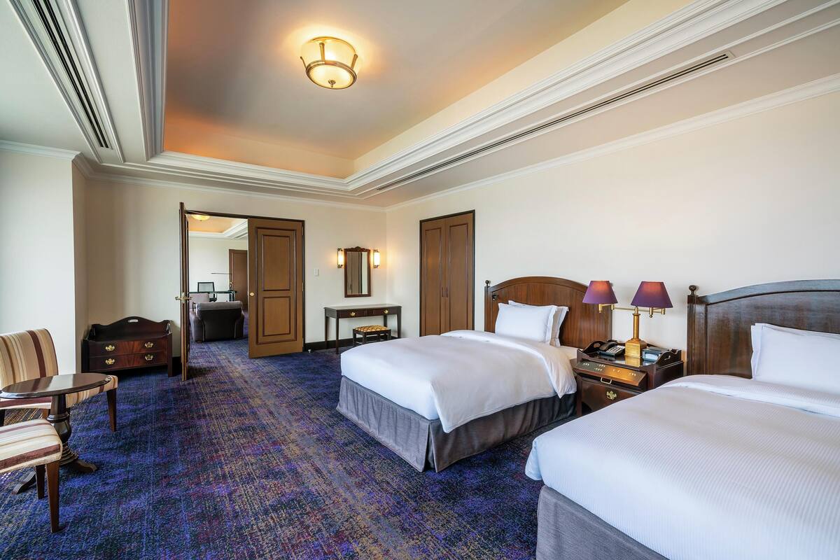 Une chambre d’hôtel avec deux lits, un bureau, une chaise, une commode et un sol recouvert de moquette.
