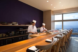 A modern restaurant with a long wooden counter, chefs in white uniforms, and a view of the city through large windows.