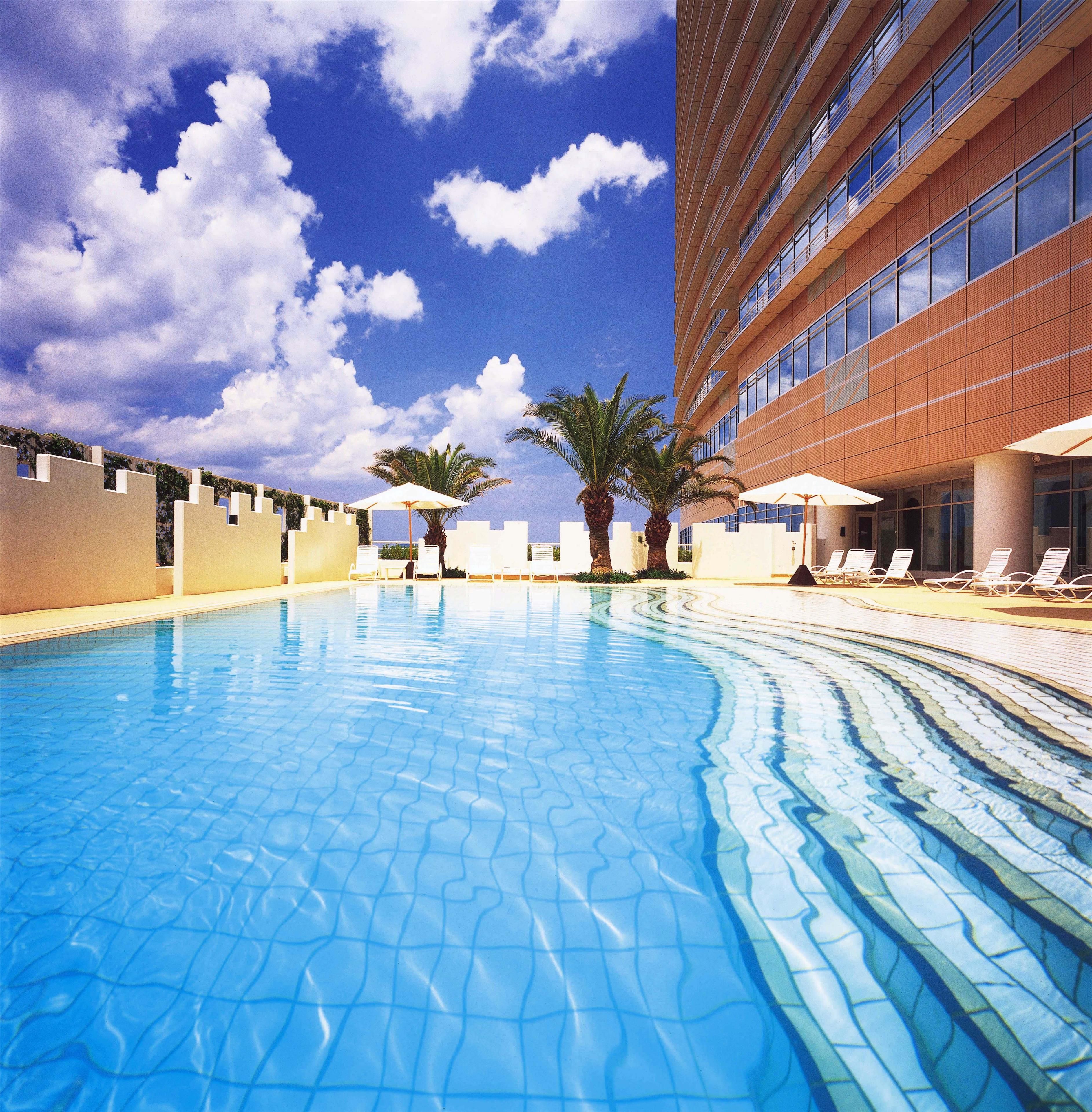 A swimming pool with lounge chairs and umbrellas, a building with a modern design, and palm trees.