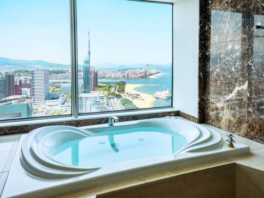 A modern bathroom with a large bathtub, a view of a cityscape through a large window, and a marble wall.