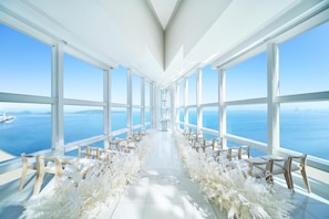 A spacious indoor venue with a long aisle flanked by chairs, large windows offering a sea view, and a white floral arrangement on the floor.