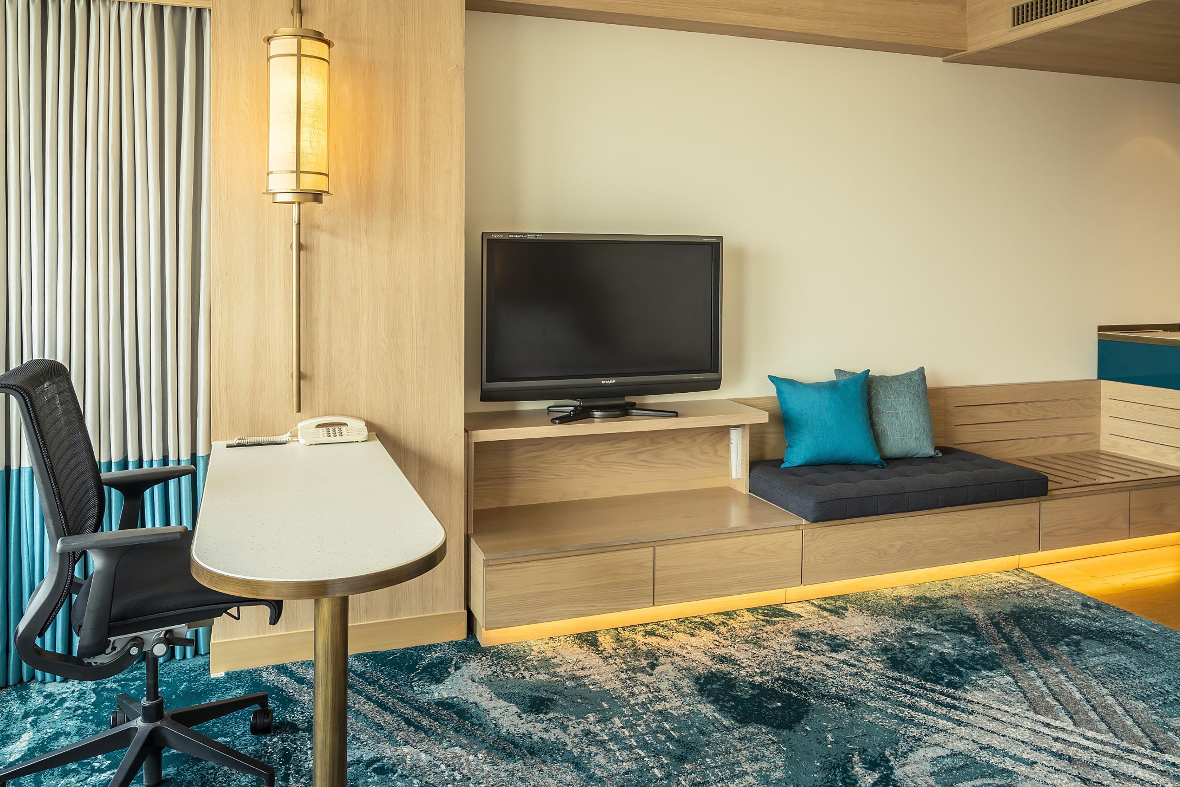 A hotel room with a desk, chair, television, and a sofa with cushions.