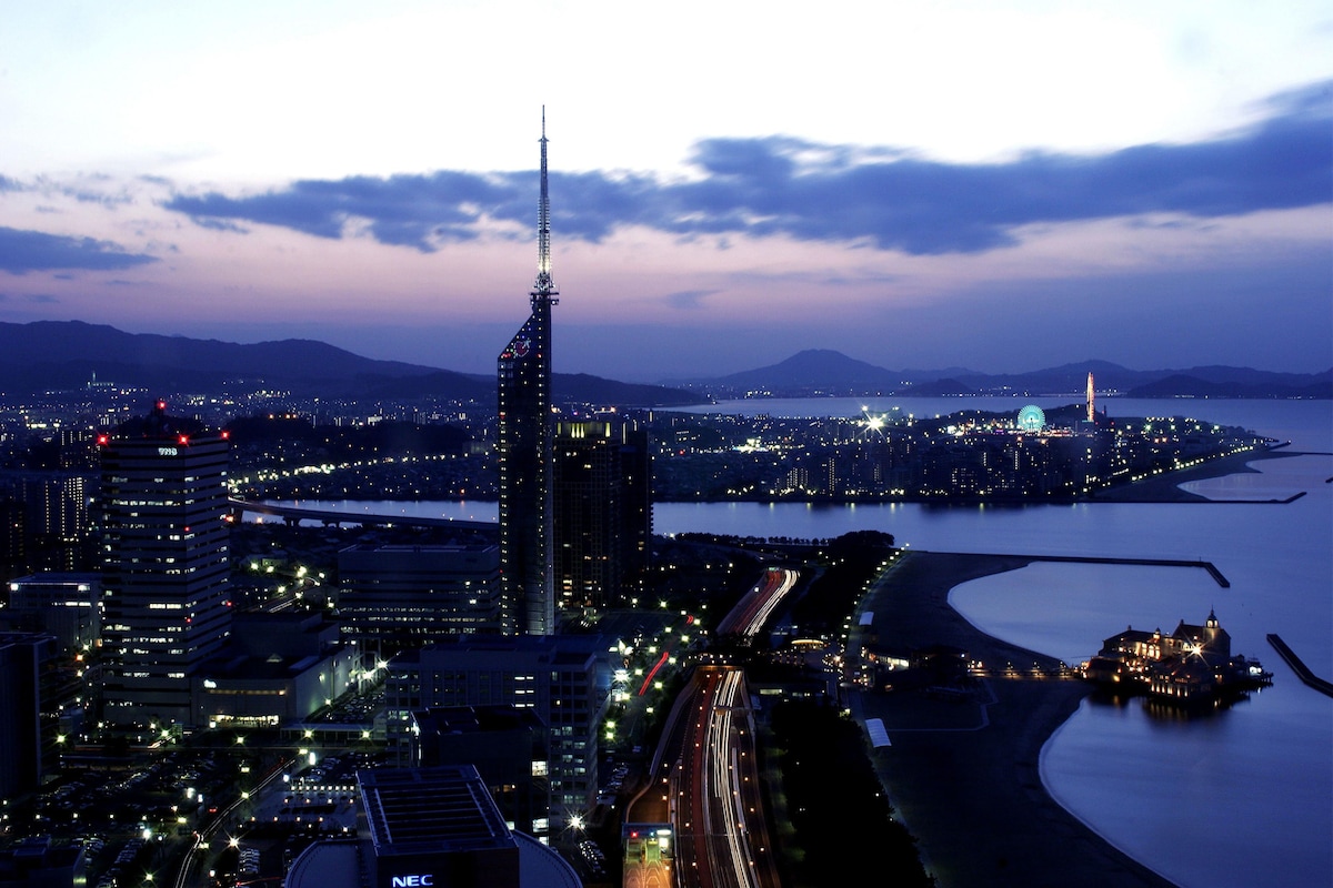 A cityscape at dusk with a prominent skyscraper, a waterfront, and a bridge.