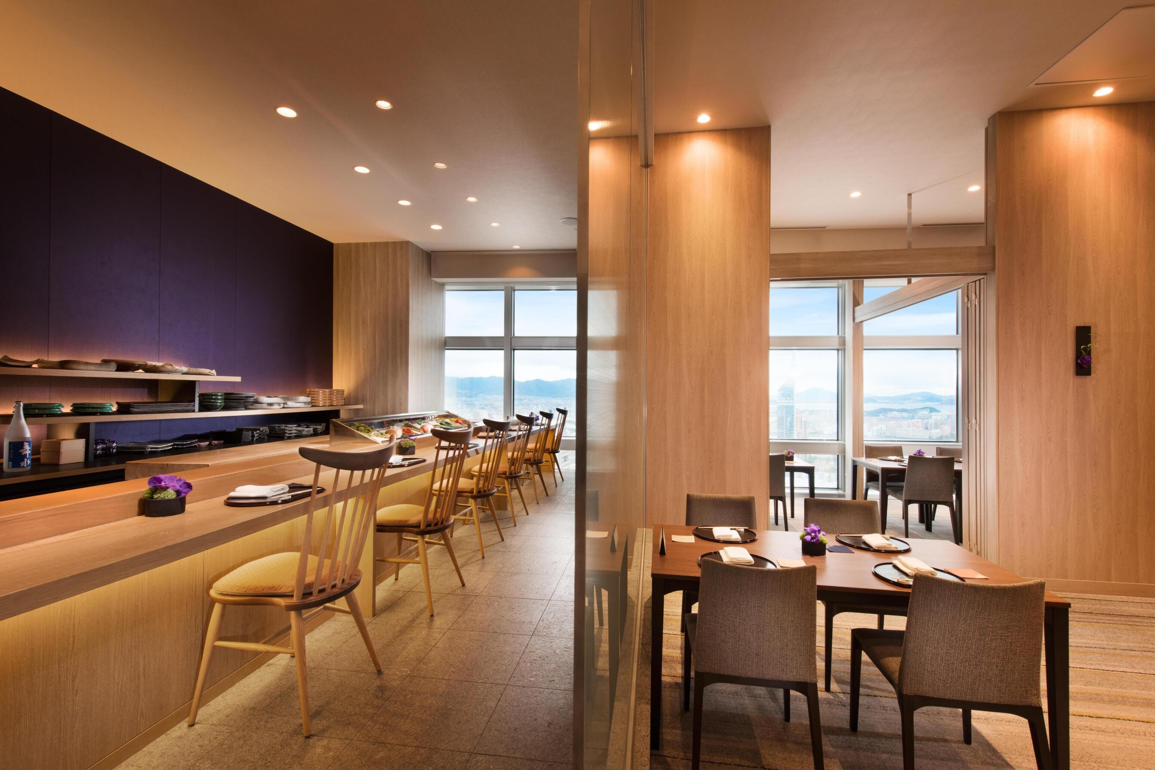 En modern hotellrestaurang med en lång bardisk, flera stolar och stora fönster med utsikt över staden.
