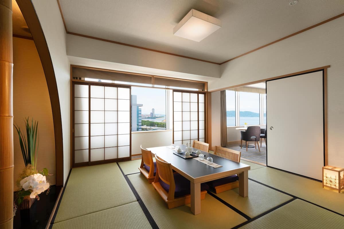 Une pièce de style japonais traditionnel, avec un sol recouvert de tatamis, une table basse, des chaises et une porte coulissante donnant sur un balcon avec vue sur la ville.