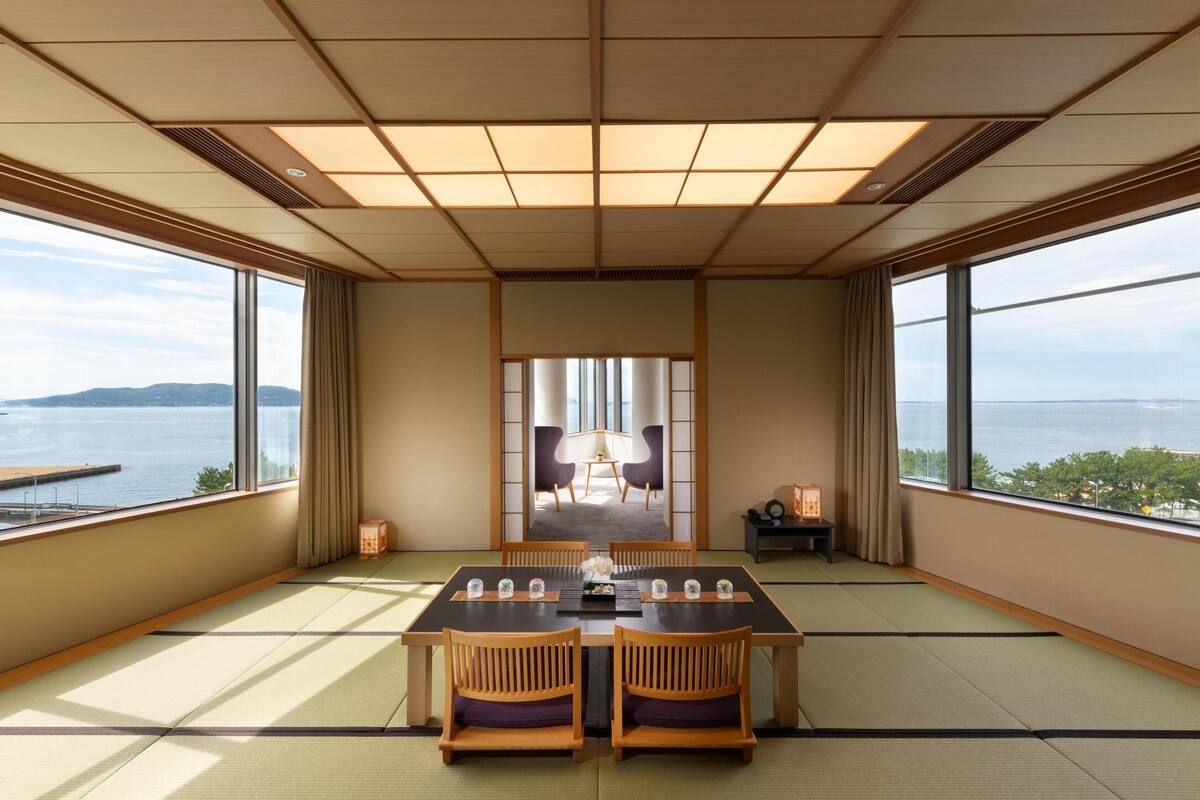Une pièce de style japonais traditionnel, avec un sol recouvert de tatamis, une table basse et des chaises, ainsi que de grandes fenêtres offrant une vue sur l’eau et les collines.