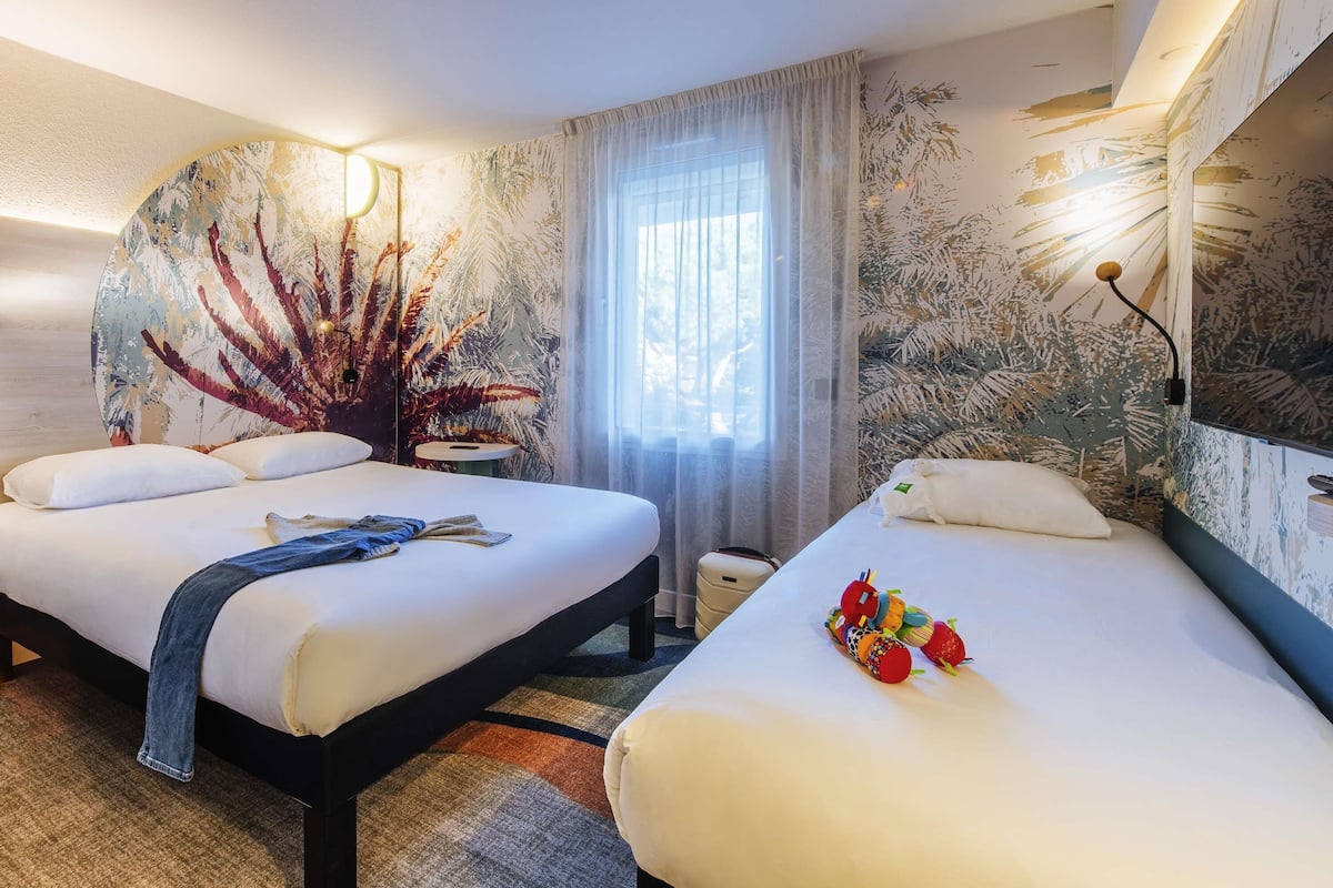 A hotel room with two beds, a large window with sheer curtains, and a wall with a leaf pattern.