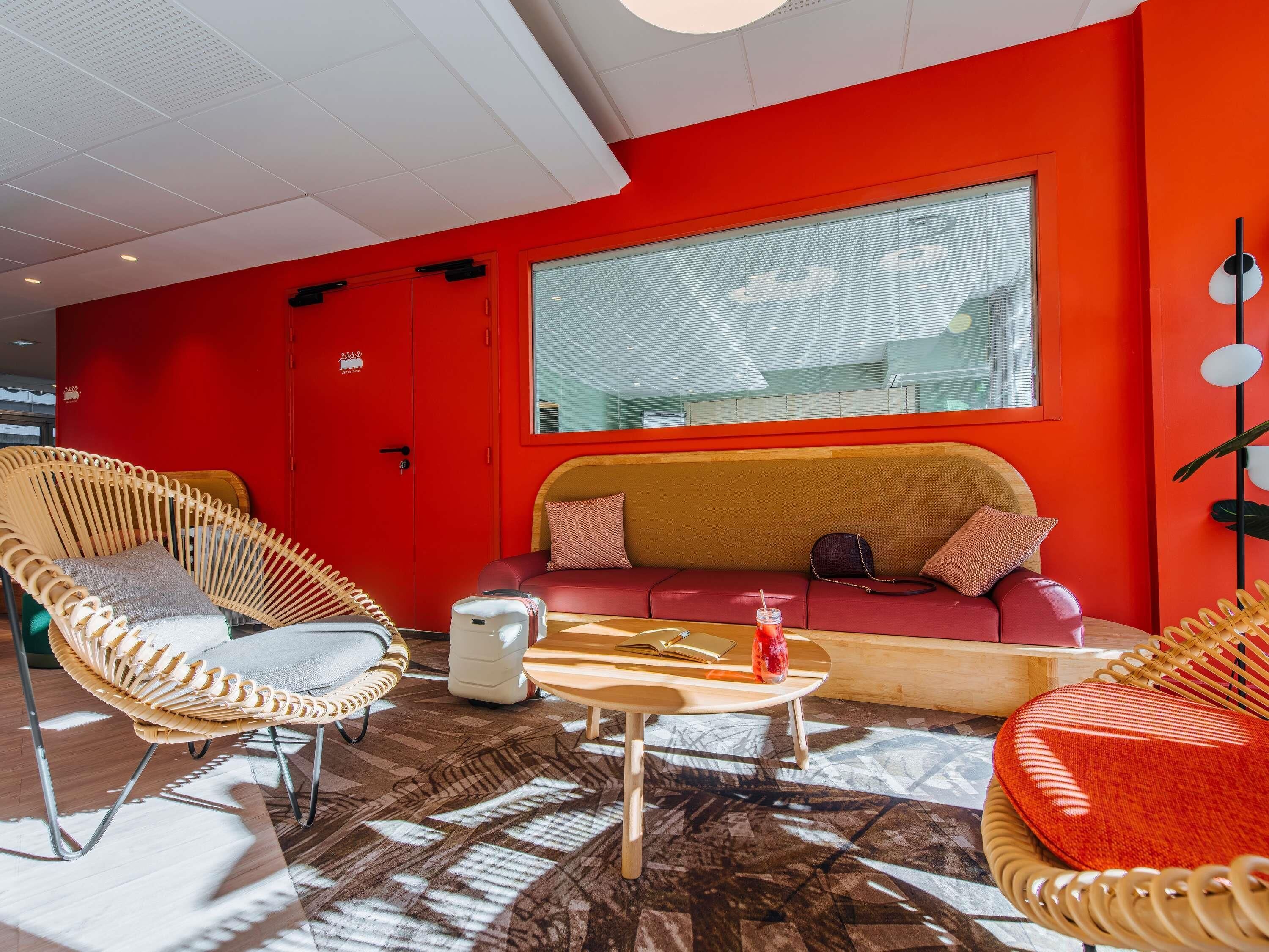 A modern room with a red wall, a large window, a sofa, two wicker chairs, a small table, and a floor lamp.