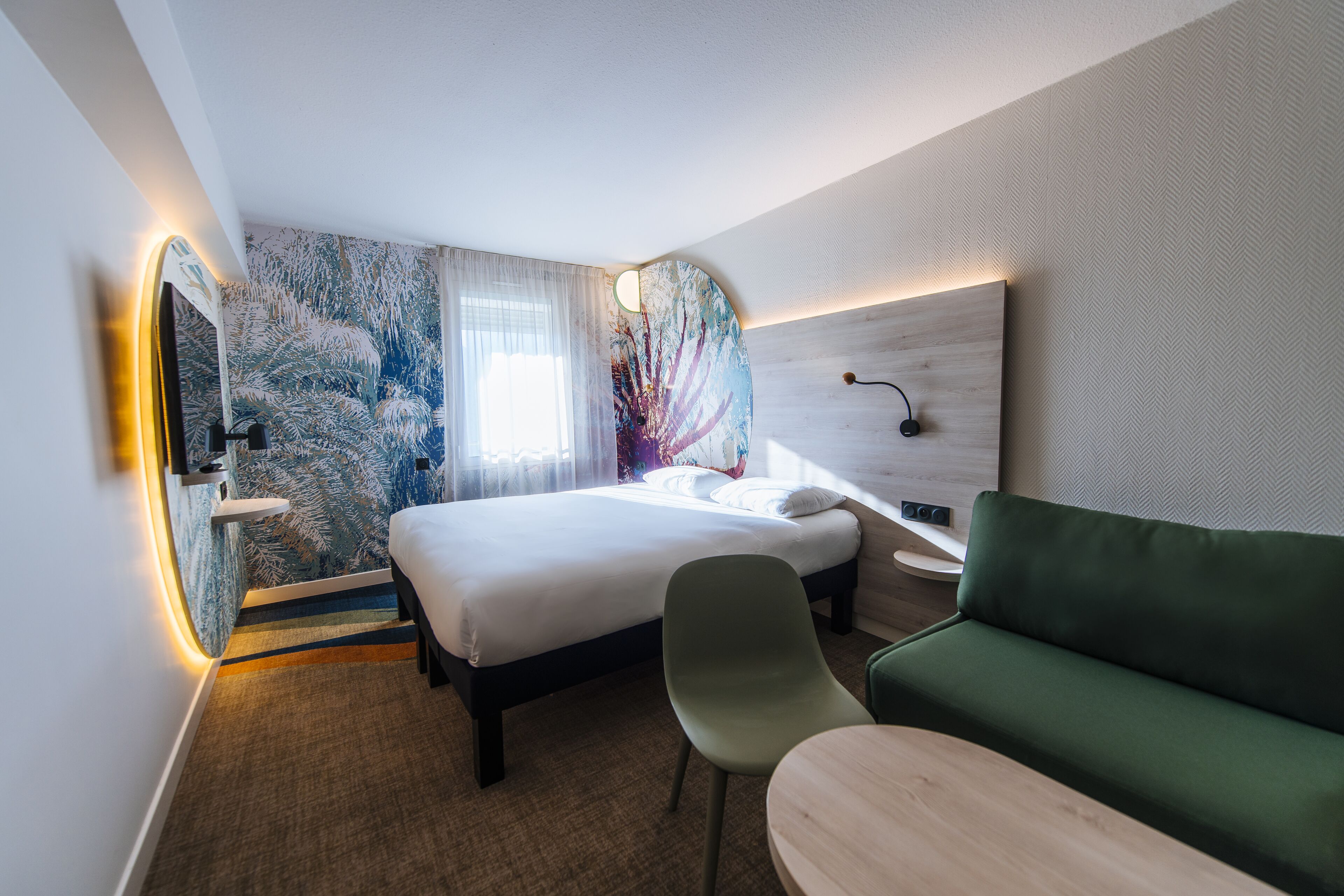 A modern hotel room with a bed, a green sofa, a small table, and a wall with a unique artwork.