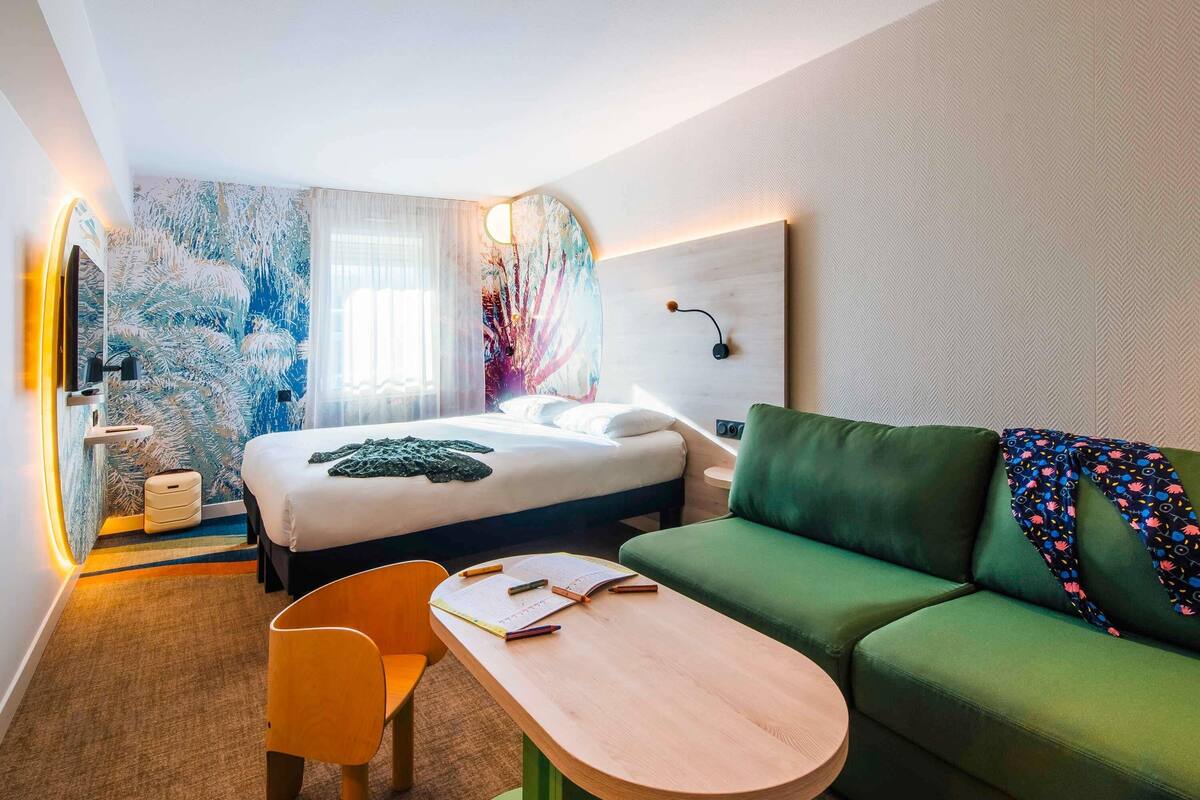 A modern hotel room with a bed, a green sofa, a wooden table, and a chair.