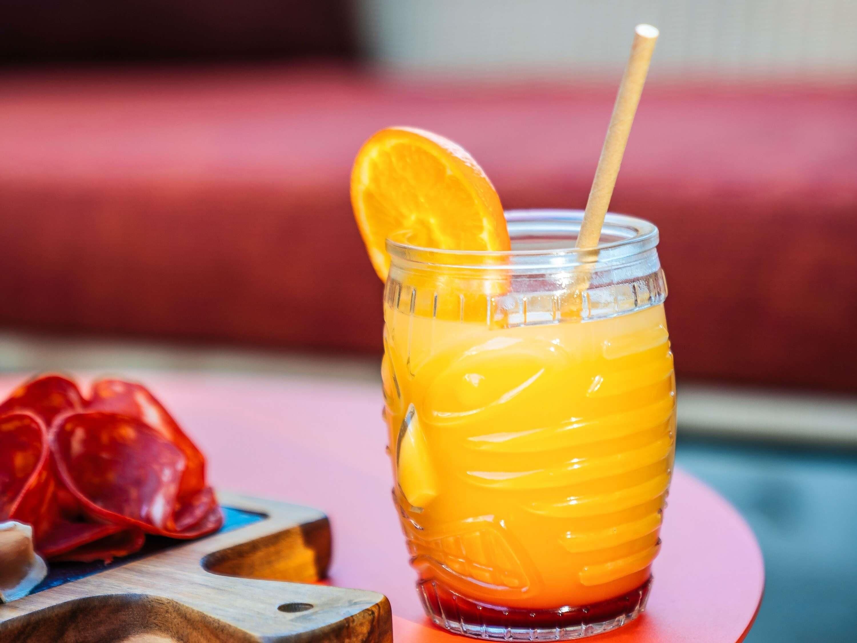 Een glas met een geel-oranje drank, een houten lepel en een sinaasappelschijfje als garnering op een rode ondergrond.