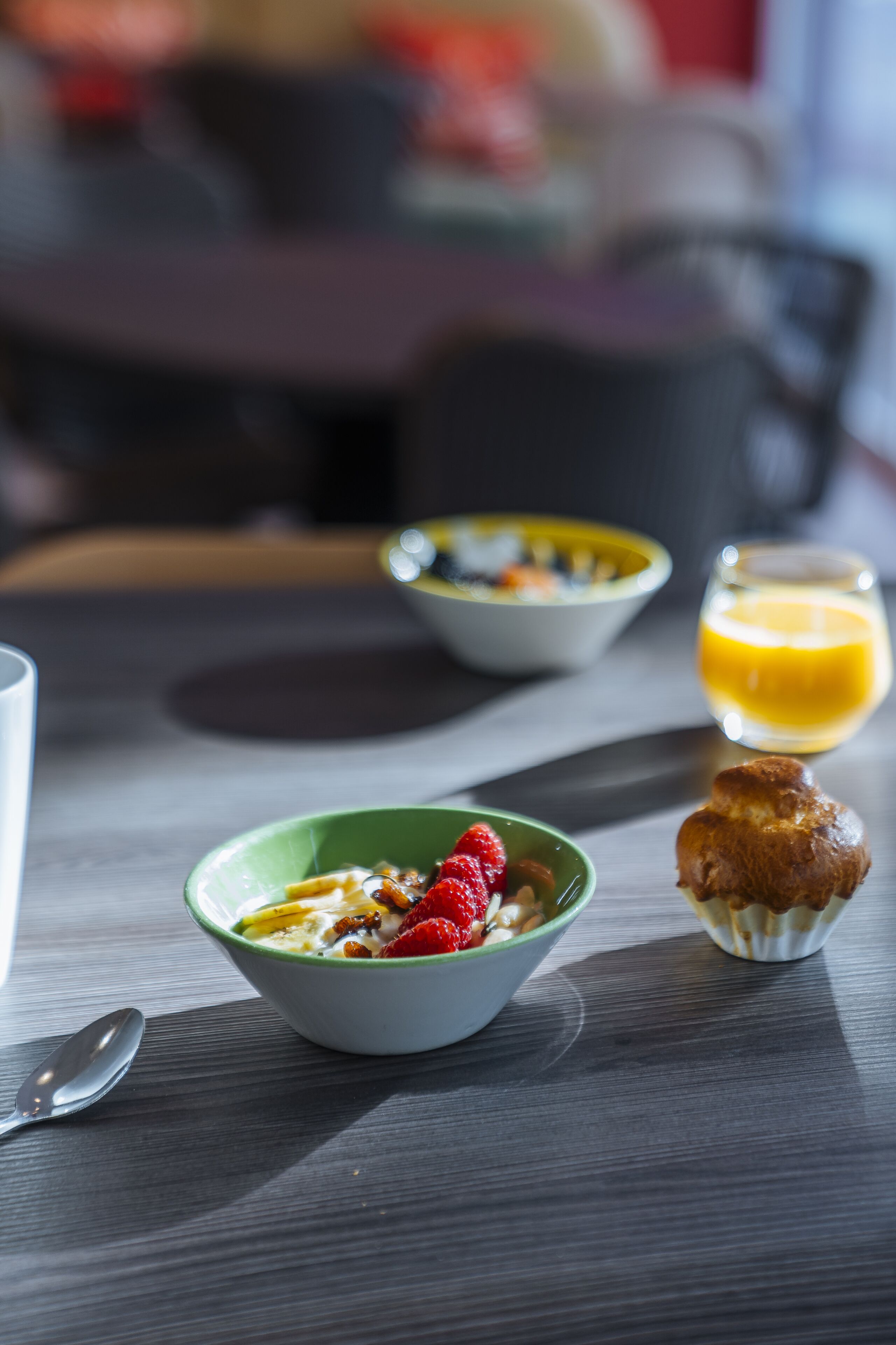 Une table mise pour le petit-déjeuner, avec des bols de céréales et des fruits, un muffin et un verre de jus d’orange.