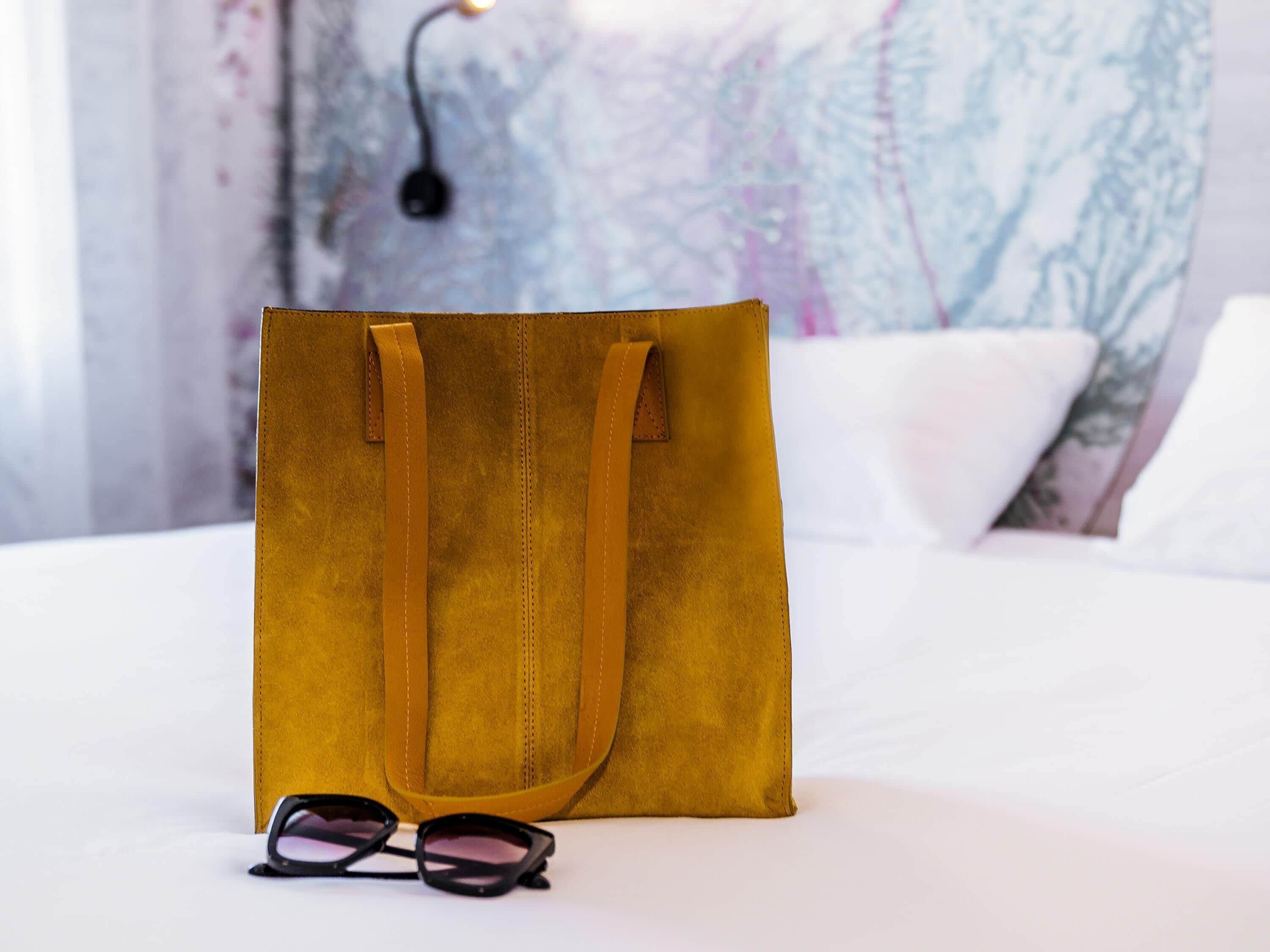 A mustard-colored tote bag on a bed with a pair of glasses beside it.