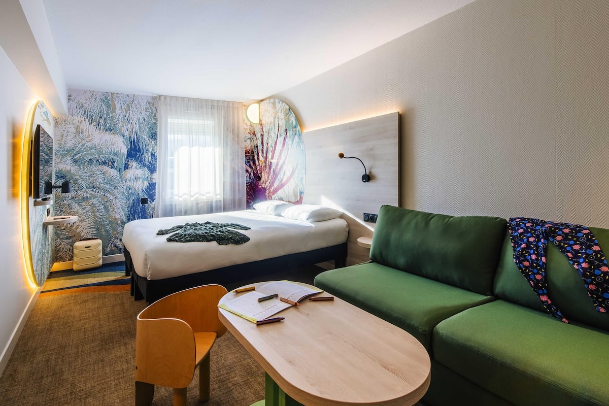 Quarto de hotel com uma cama, um sofá verde, uma mesa de madeira, uma cadeira e uma parede com luminária.