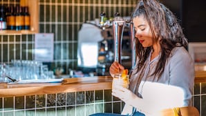 Una persona sentada en la barra con una bebida en la mano, un menú sobre el mostrador y botellas de alcohol al fondo.