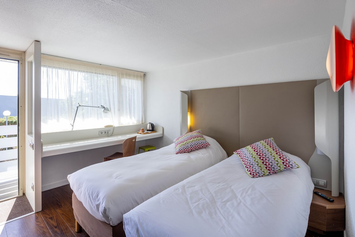 A hotel room with two beds, a desk with a laptop, a chair, and a window with curtains.
