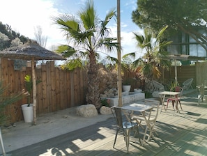 A patio area with a wooden fence, palm trees, a thatched roof structure, and outdoor furniture. - Noemys Arles (Arles)